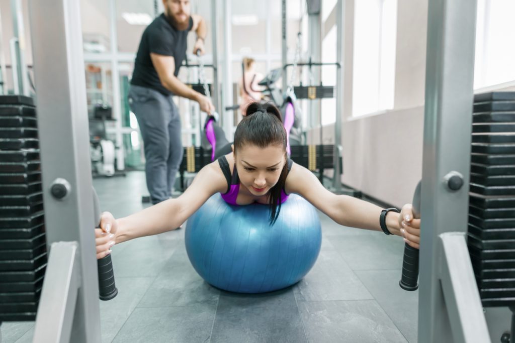 Kinesis technology, kinesitherapy, healthy lifestyle. Young woman doing rehabilitation exercises with personal instructor using kinesi machine, fitness gym background.