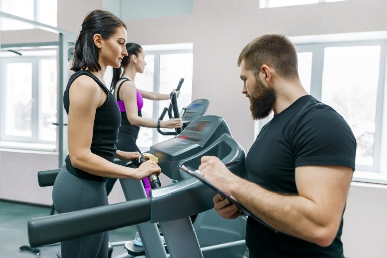 Young smiling fitness women with personal trainer an adult athletic man on treadmill in the gym. Sport, teamwork, training, healthy lifestyle concept.