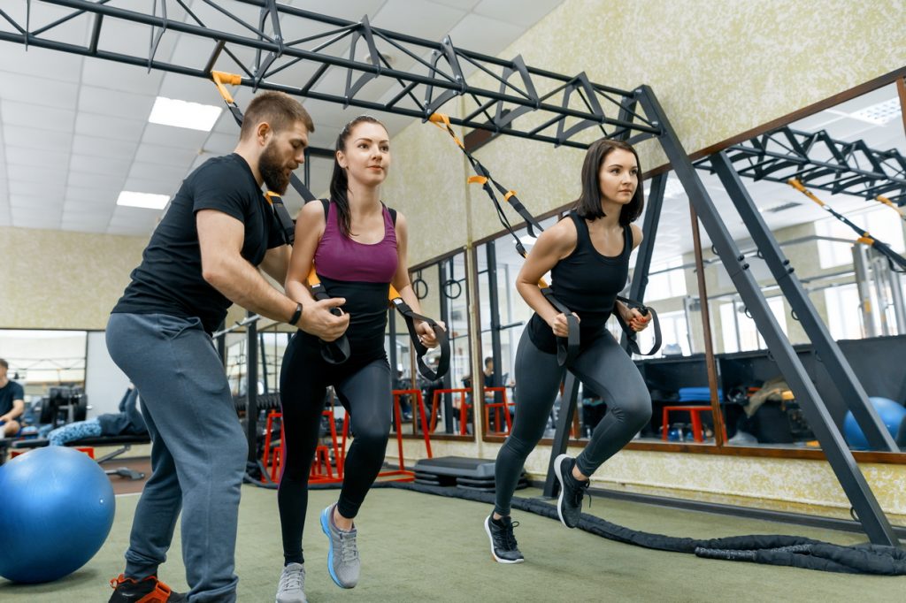 Fitness trainer coaching and helping women doing exercises on cross fit using straps system in gym. Sport, teamwork, training, people concept
