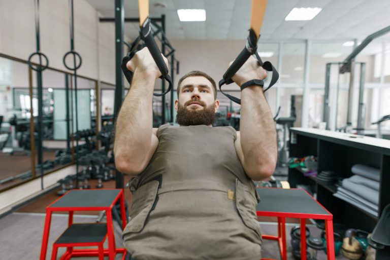 Military sport, muscular caucasian bearded adult man doing exercises in the gym dressed in a bulletproof armored vest.
