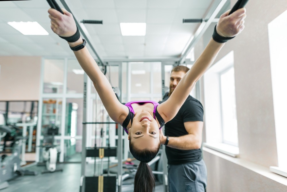 Kinesis technology, kinesitherapy, healthy lifestyle. Young woman doing rehabilitation exercises with personal instructor using kinesi machine, fitness gym background.